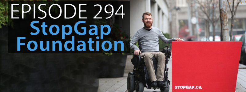 Luke Anderson, dressed in a grey sweater and tan pants, sits in his wheelchair posed beside a portable wooden ramp that stands upright emblazoned with the words stopgap.ca.